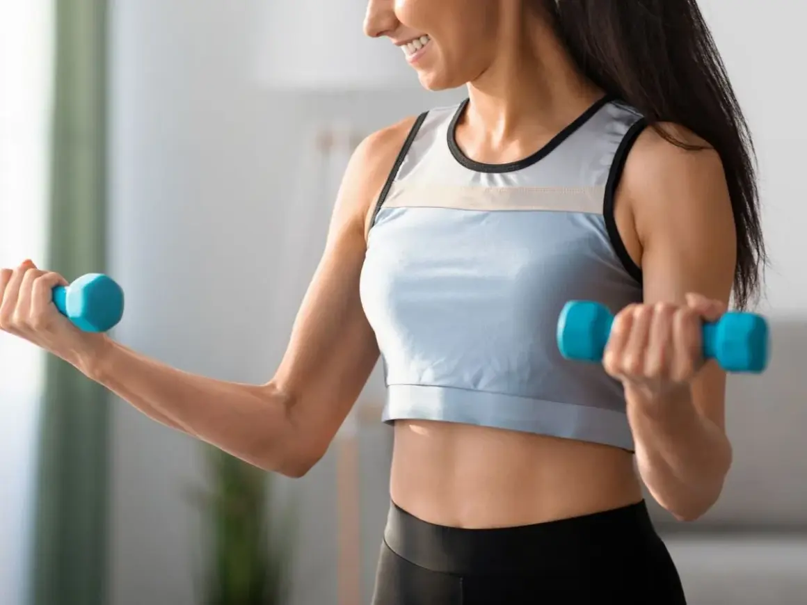 Uśmiechnięta kobieta wykonuje ćwiczenia na biceps w domu, podnosząc niebieskie hantle.