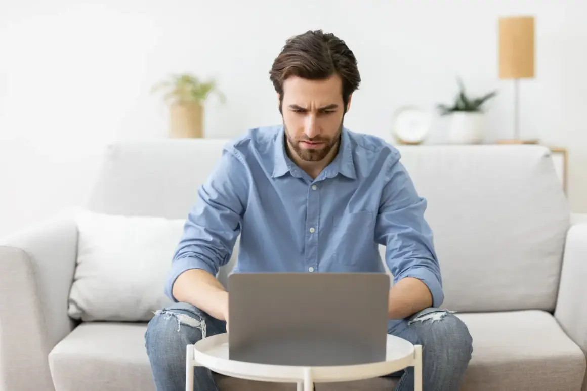 A man with a beard, wearing a blue shirt and ripped jeans, looks intently at a laptop on a small table.
