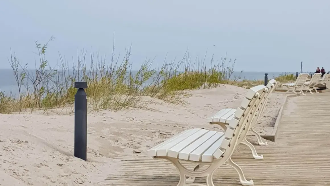 Białe ławki na drewnianej promenadzie w Dziwnowie, z widokiem na piaszczyste wydmy i morze.