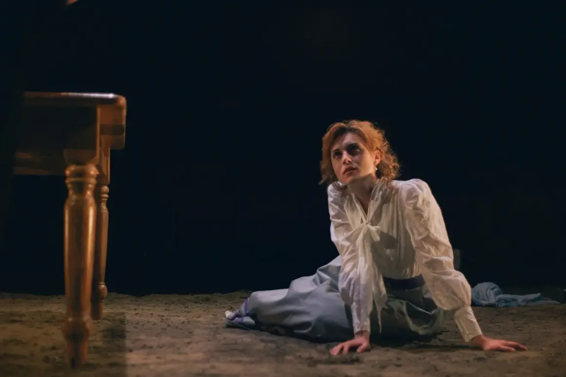 La signorina Julie, con capelli rossi e un vestito bianco, è inginocchiata su un pavimento di terra, con uno sguardo perso nel vuoto.