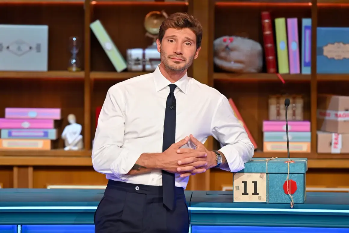 Stefano De Martino outfit: camicia bianca, cravatta blu scuro e pantaloni eleganti. Sullo sfondo, una libreria piena di oggetti colorati.