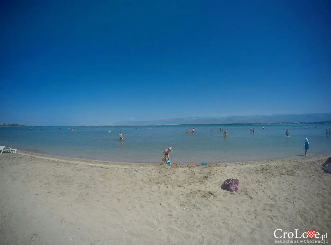 Piaszczysta plaża w Chorwacji z ludźmi kąpiącymi się w morzu i dzieckiem bawiącym się na piasku.