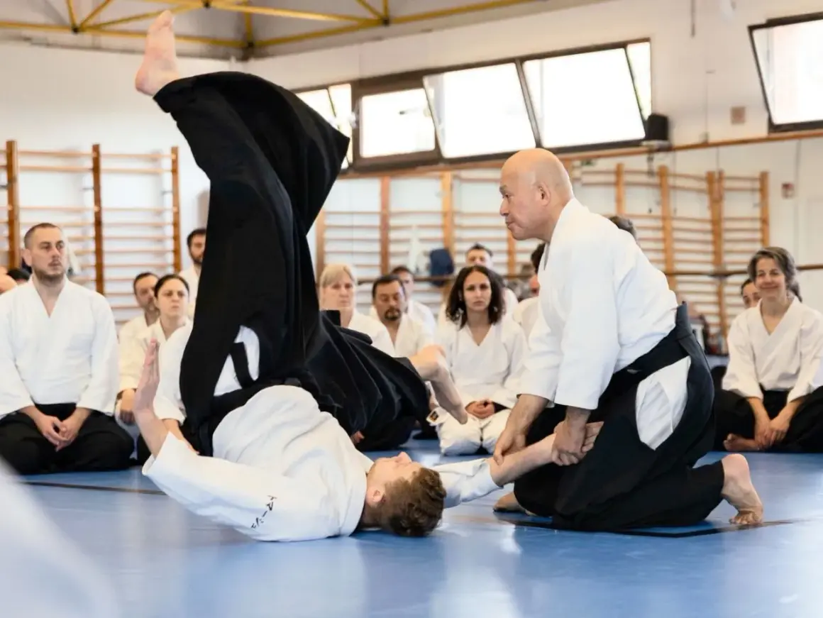 Två personer i vit gi utför en kastteknik inom kampsporten aikido. Åskådare sitter på golvet och observerar.