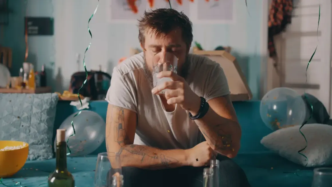 Zmęczony mężczyzna z tatuażami pije wodę po imprezie, symbolizującą jego odstawienie alkoholu. W tle resztki zabawy.
