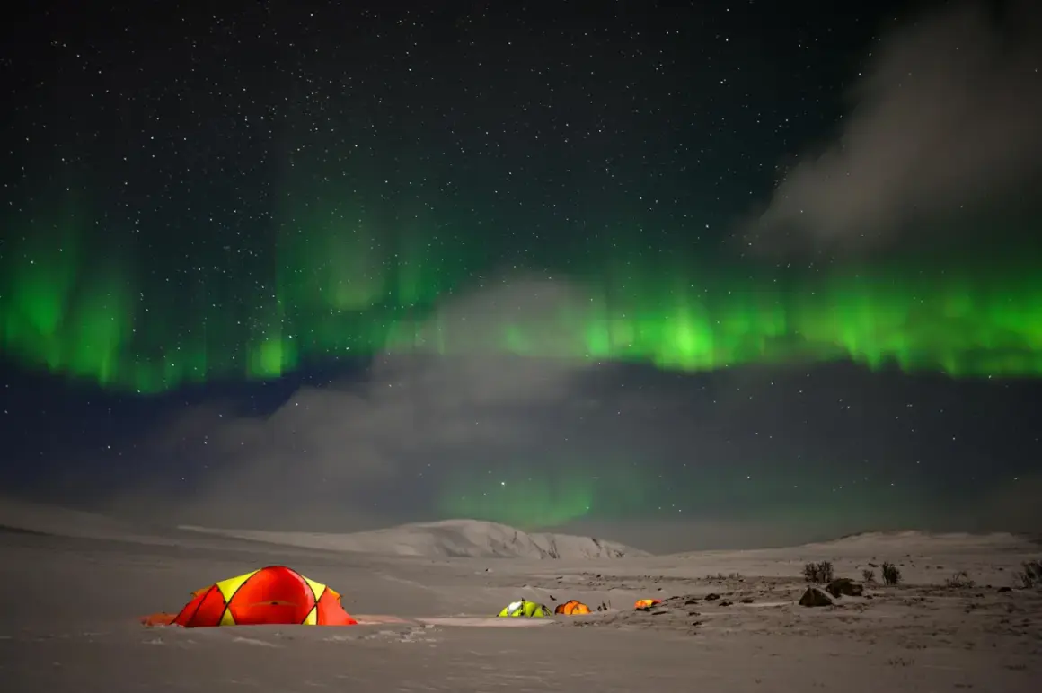 Nocne niebo nad Norwegią rozświetla zorza polarna. Kilka kolorowych namiotów rozbitych na śniegu tworzy przytulne schronienie pod tym niezwykłym zjawiskiem.