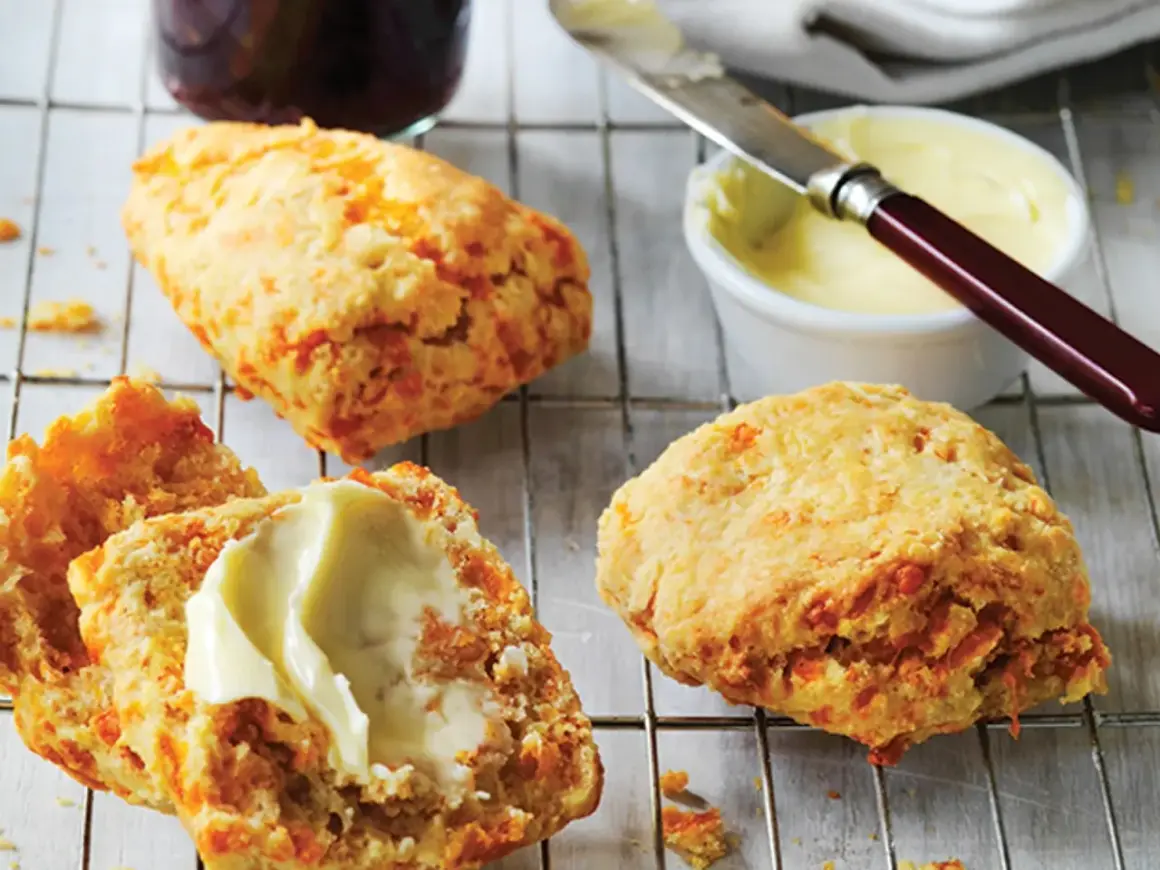 Trois scones américains dorés, dont un coupé en deux et garni de beurre, sur une grille. Un pot de confiture et du beurre à tartiner sont à proximité.