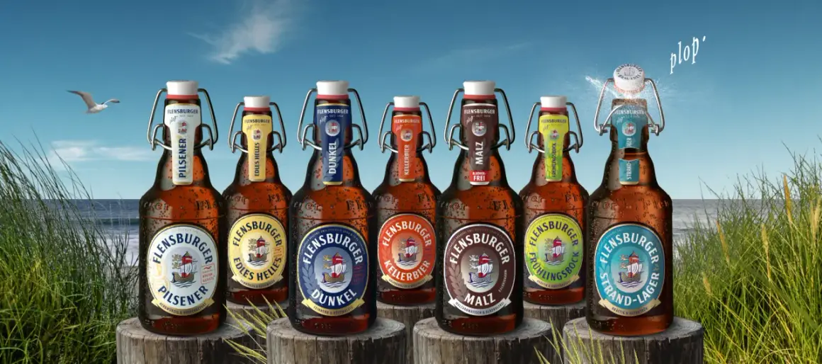 Flensburger Pils Bier Marken am Strand. Eine Möwe fliegt über das Meer, während die Flaschen mit Bügelverschluss im Gras stehen.