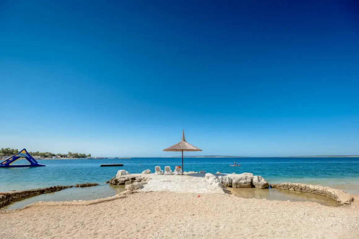 Słoneczna plaża z parasolem i leżakami w camping village Simuni. W tle wodny plac zabaw i spokojne morze.
