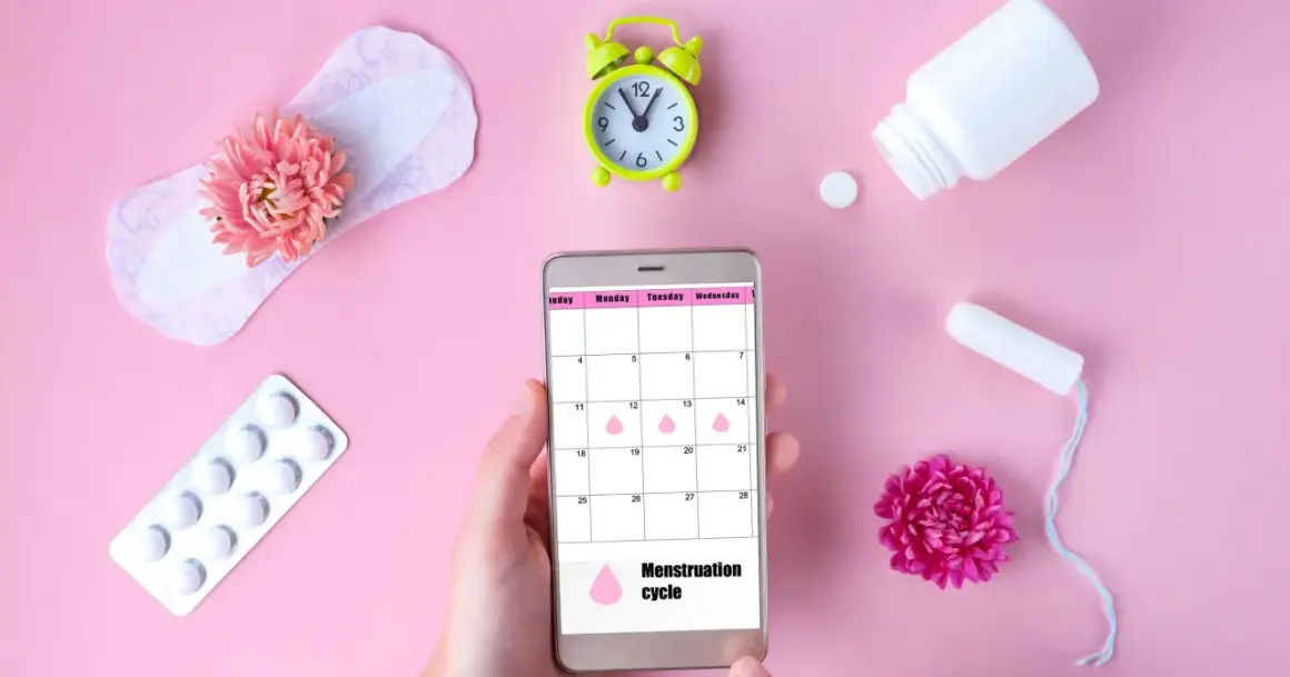 Telefon z kalendarzem cyklu menstruacyjnego, obok podpaska, tampon, leki i kwiaty. Warto pamiętać o mięśniakach macicy.