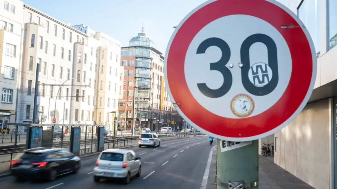 Verkehrsschild "Tempo 30 Berlin" auf einer Straße mit Autos und Gebäuden im Hintergrund.
