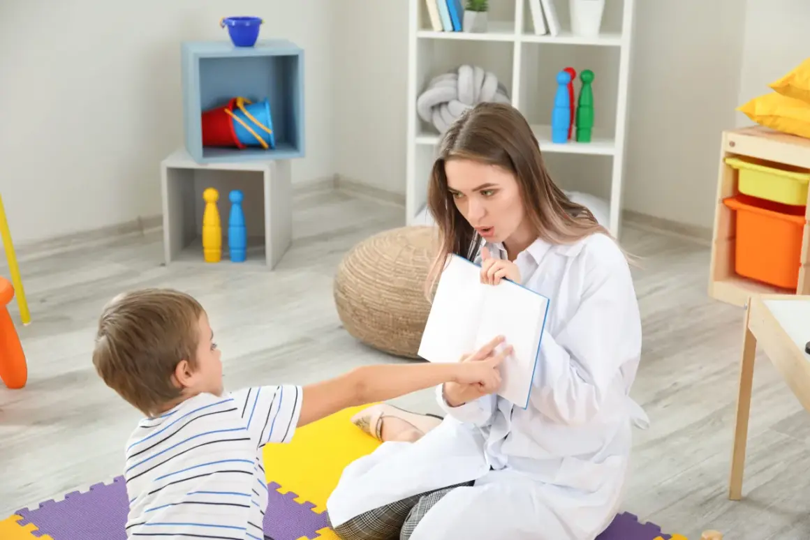 Terapia logopedyczna z dzieckiem z opóźnionym rozwojem mowy. Terapeuta pokazuje dziecku książkę, ćwicząc wymowę.