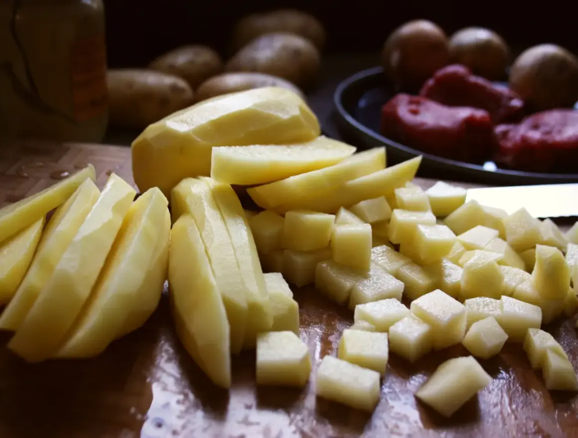 Skalade och skurna potatisar i olika former, redo för att bli härligt råstekt potatis. I bakgrunden syns hela potatisar och kött.