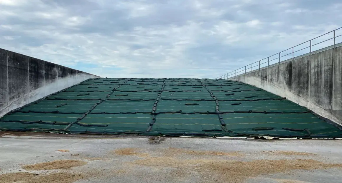 Ampia trincea agricola coperta da telo verde, con pareti in cemento e cielo nuvoloso.