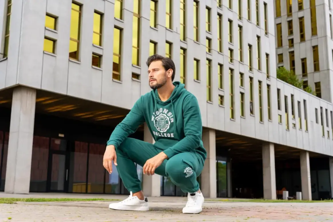 Mężczyzna w dresie butelkowa zieleń z nadrukiem "College" kuca przed nowoczesnym budynkiem z żółtymi oknami.