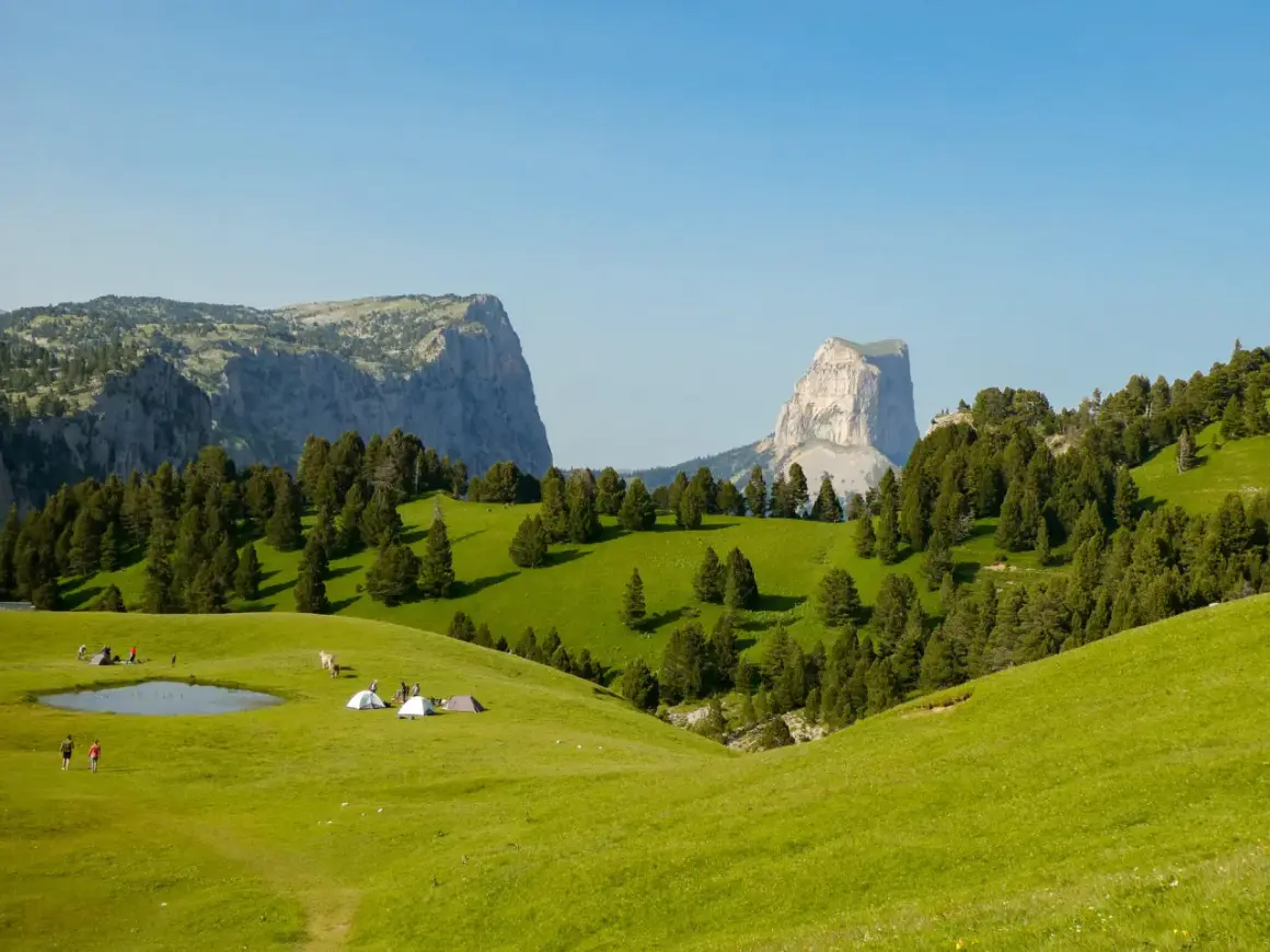 Un campement de tentes et de randonneurs sur une prairie verdoyante, avec des montagnes imposantes en arrière-plan. Le **pas de l'aiguille** est visible au loin.