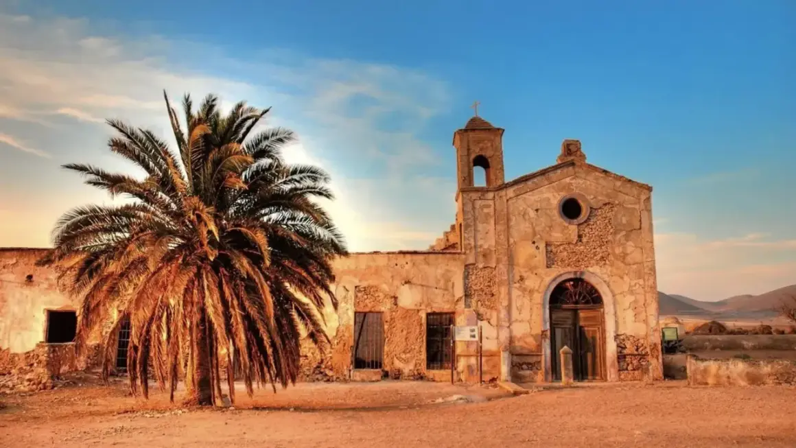 El cortijo del fraile, una edificación antigua con una palmera imponente, bajo un cielo azul y nubes al atardecer.