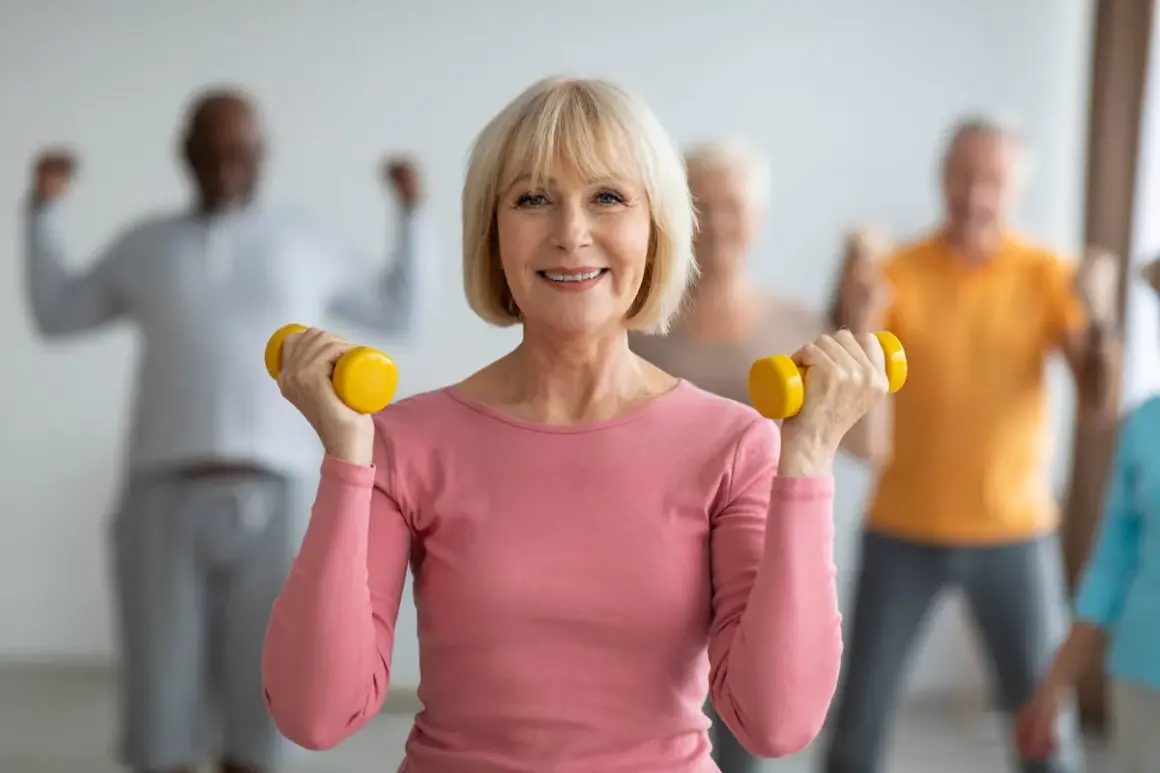 Une femme souriante fait de la gymnastique senior avec des haltères jaunes. D'autres personnes âgées s'entraînent en arrière-plan.