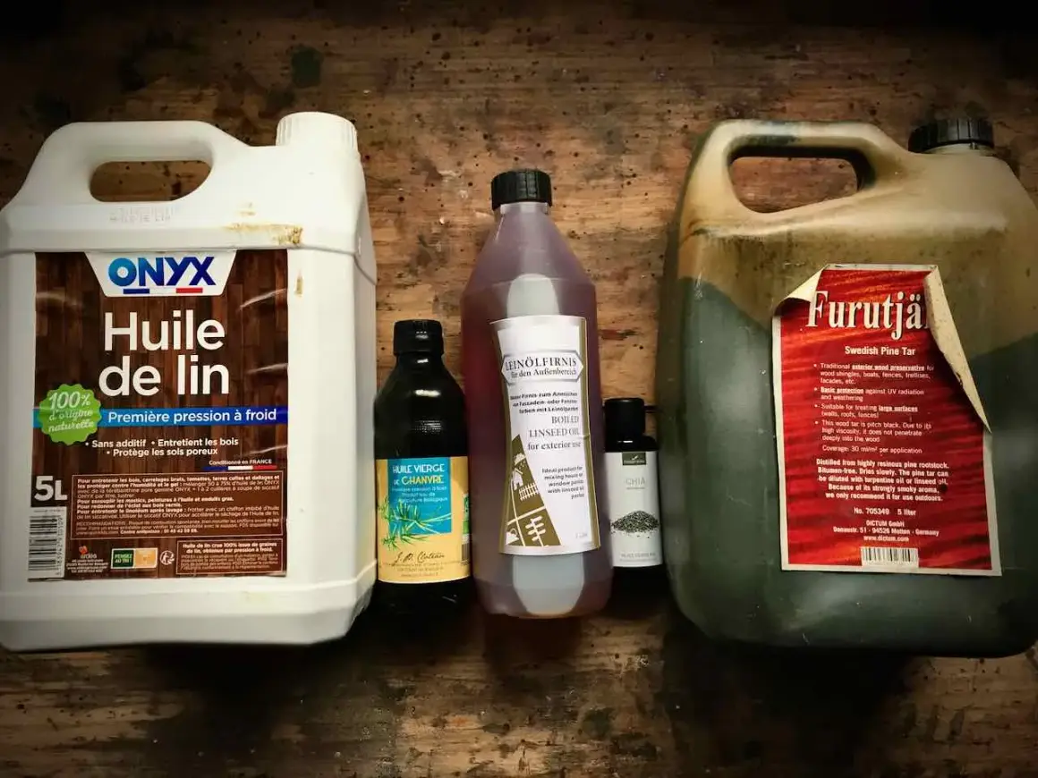 Plusieurs bouteilles d'huile pour le bois, dont de l'huile de lin et du goudron de pin, sont alignées sur une surface en bois.
