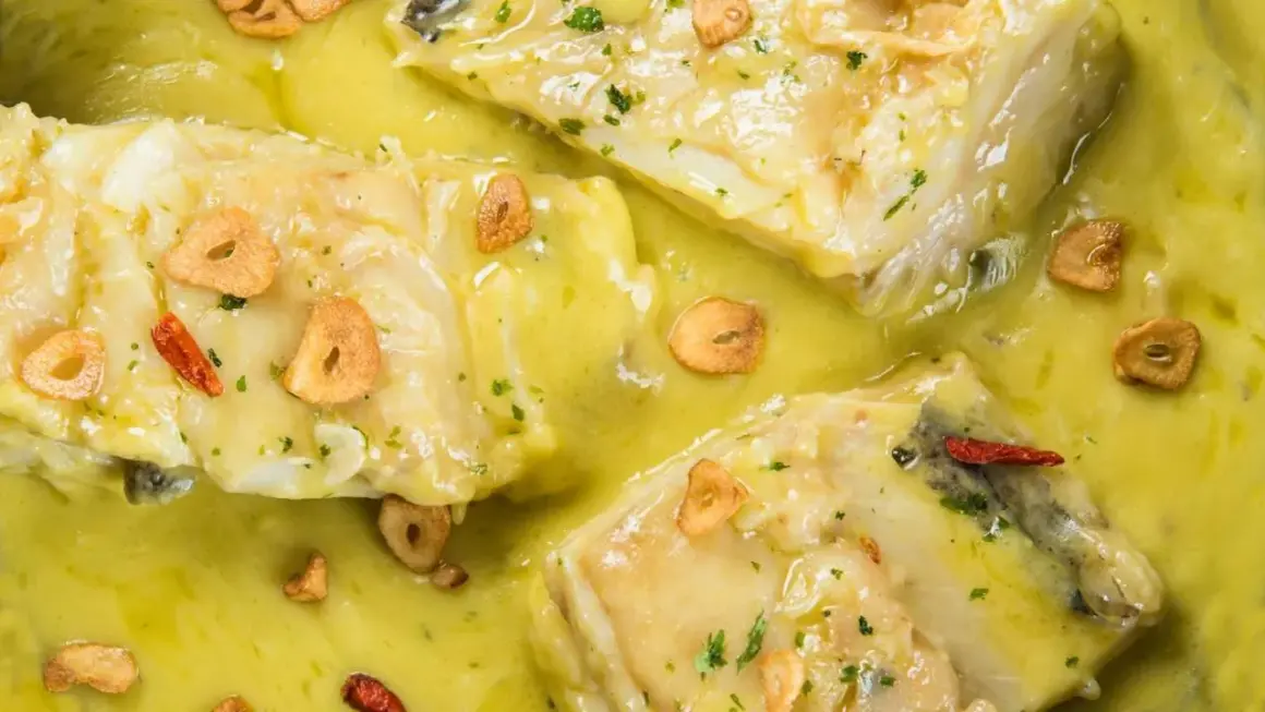 Merluza al pil pil, con trozos de pescado blanco en una salsa amarilla cremosa, adornada con láminas de ajo crujiente y guindilla.