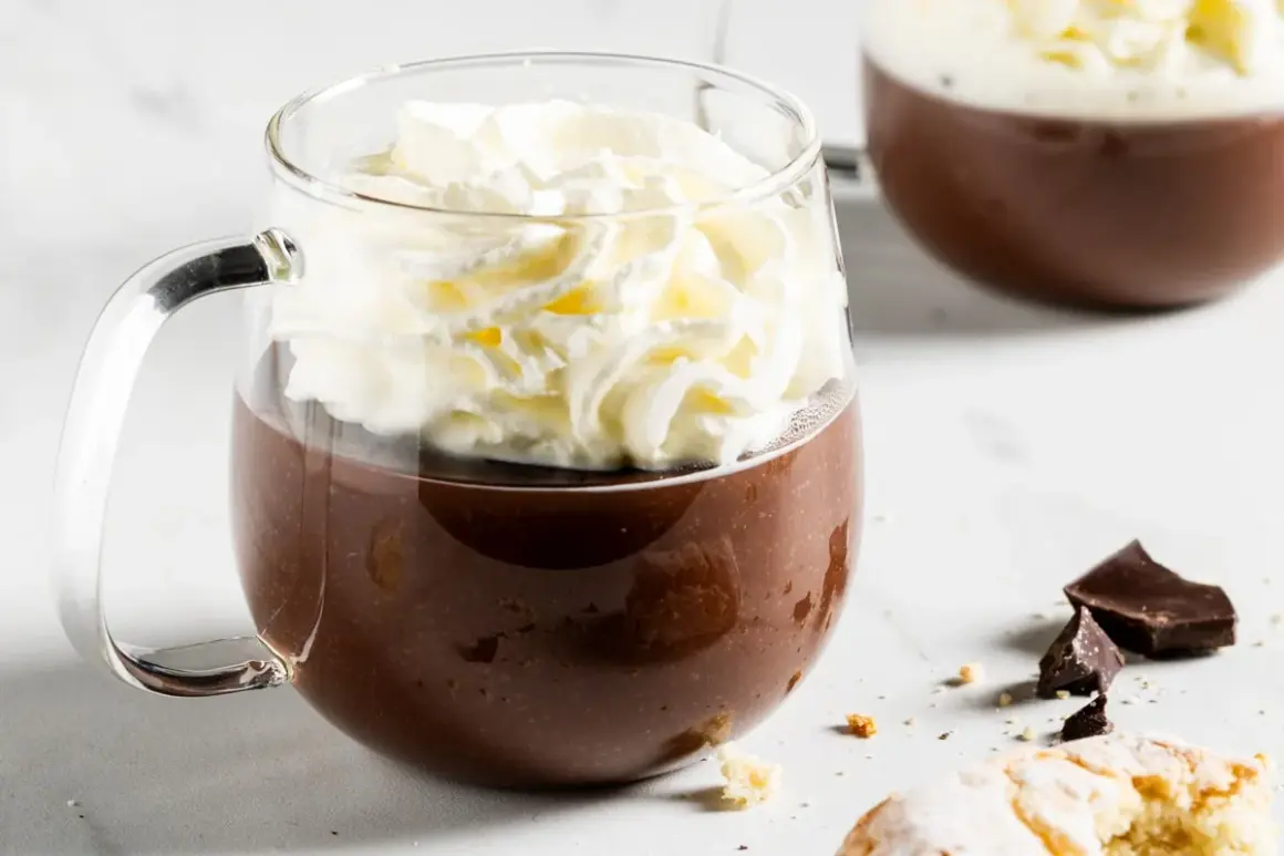 Una tazza di fumante cioccolata calda coronata da panna montata, con pezzi di cioccolato e biscotti sullo sfondo.