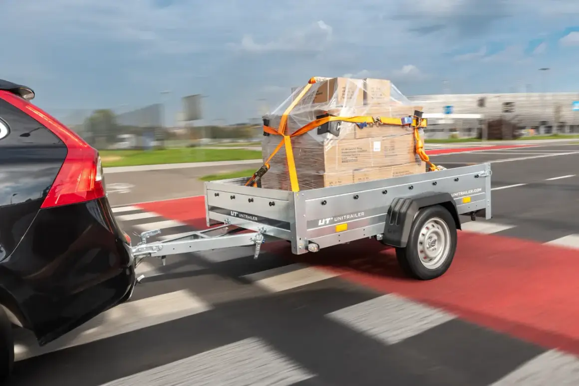 Nowy projekt przyczepki samochodowej UNITRAILER w akcji. Pudełka zabezpieczone pasami na przyczepce ciągniętej przez czarne auto.