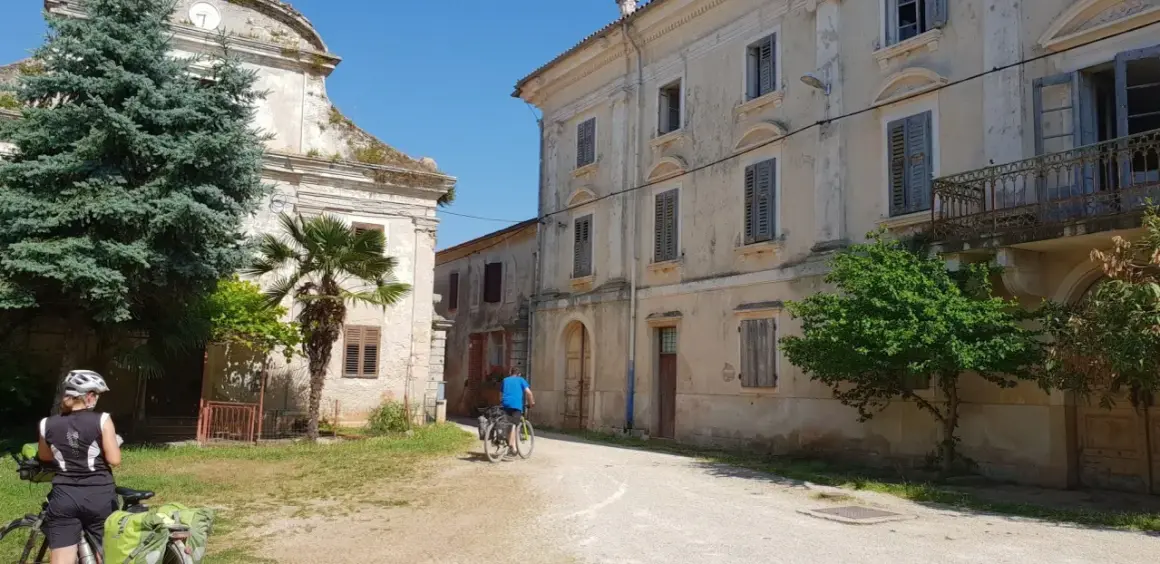 Ciclistas exploran un pueblo antiguo en Croacia, con edificios de piedra y vegetación exuberante.