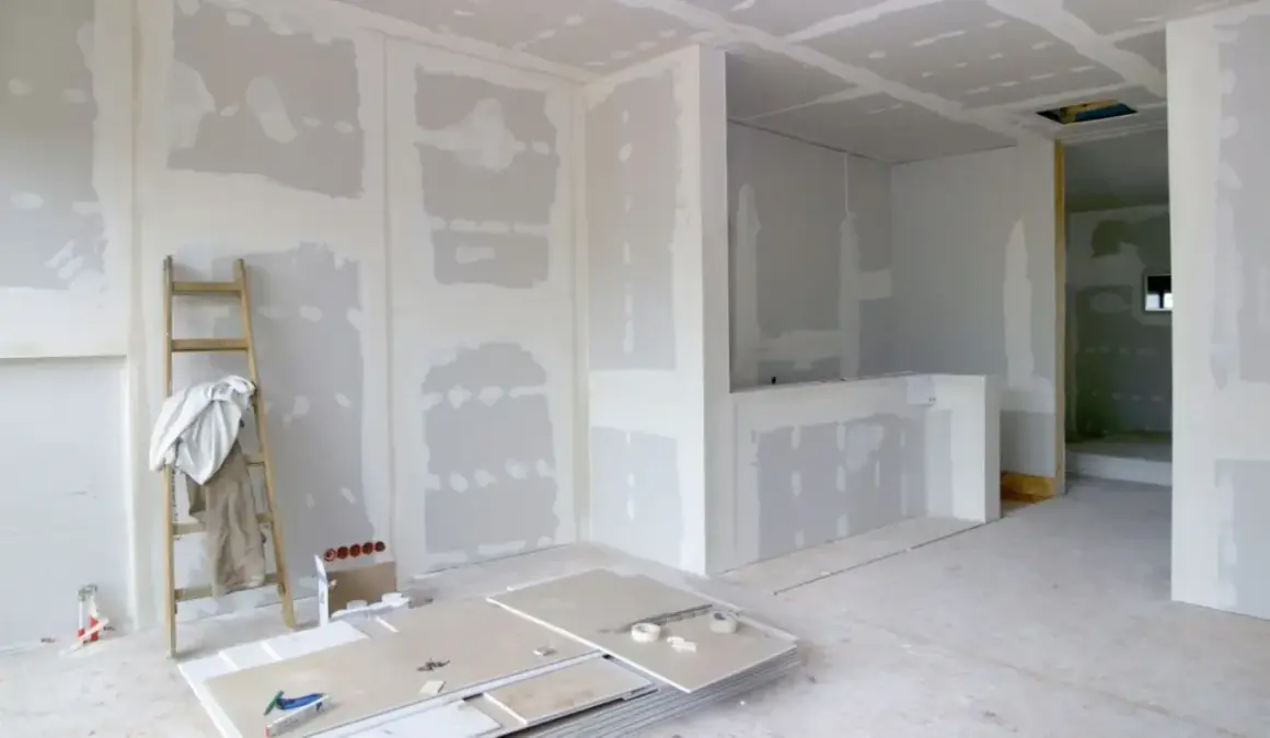 Interni in fase di finitura, con pareti in cartongesso pronte per essere stuccate. Una scala appoggiata al muro, pannelli e attrezzi a terra suggeriscono come fare un muro.
