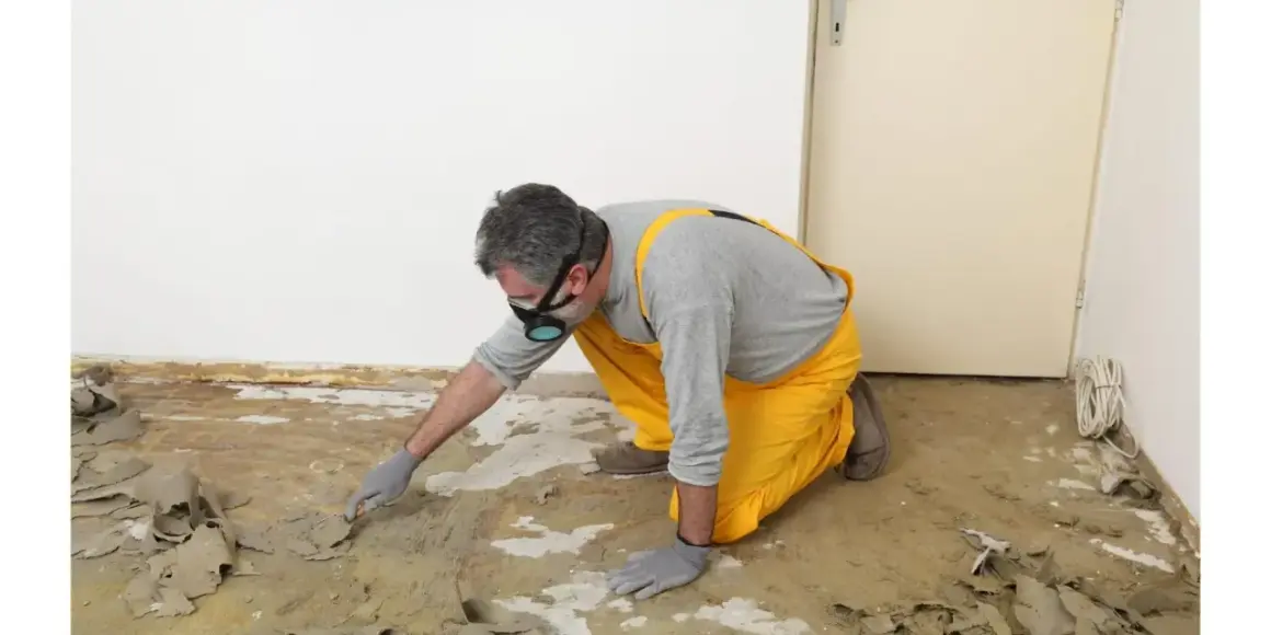 Un homme en salopette jaune et masque retire la colle de moquette avec une spatule. Une méthode naturelle pour enlever colle moquette.