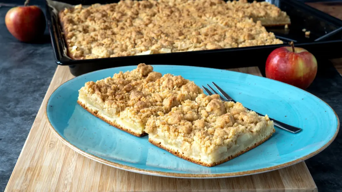 Zwei Stücke saftiger Apfelkuchen mit Streuseln auf einem blauen Teller, daneben ein ganzer Apfel. Perfekt für ein Apfelkuchen Rezept.