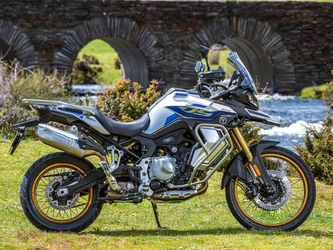 Moto Voge 500DS, una moto china con motor BMW, lista para la aventura junto a un puente de piedra y un río.