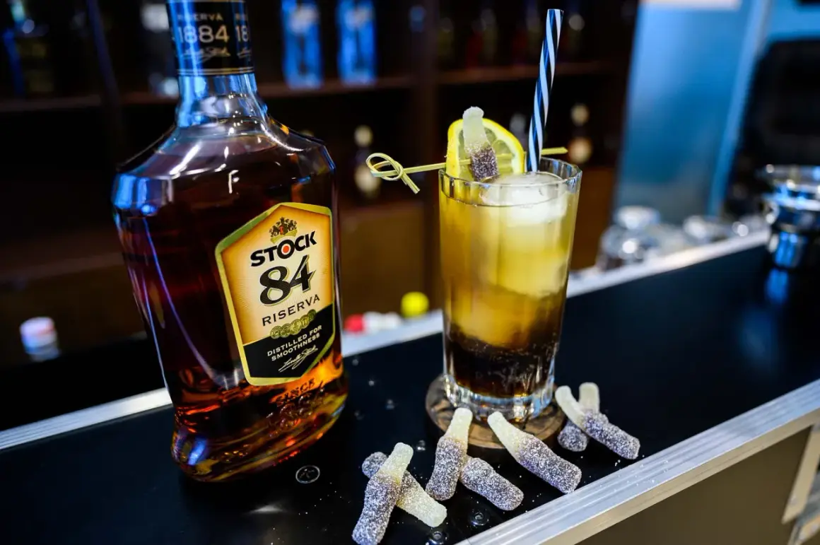 Butelka brandy Stock 84 i drink z cytryną, słomką i żelkami w kształcie butelek.
