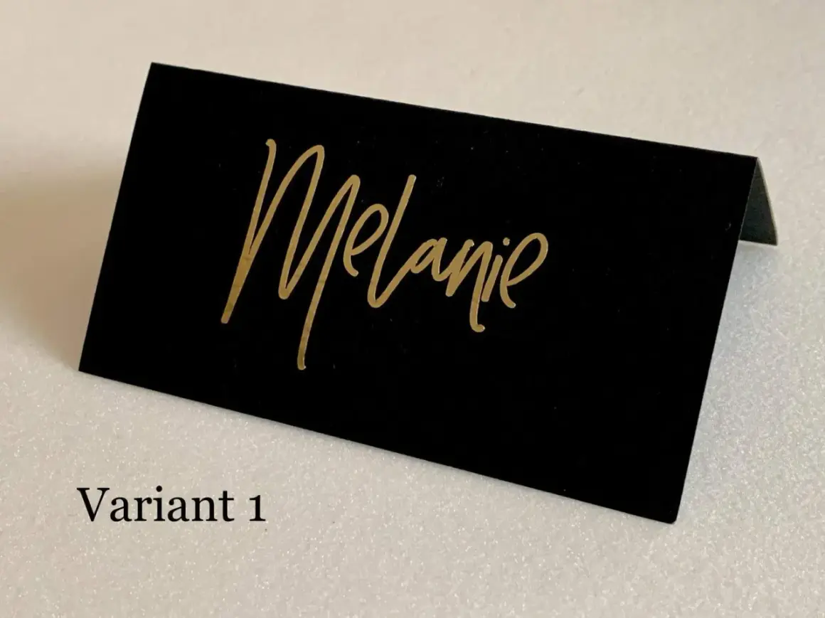Marque place noir et or avec le prénom "Melanie" écrit en calligraphie dorée. Un élégant détail pour une table de fête.