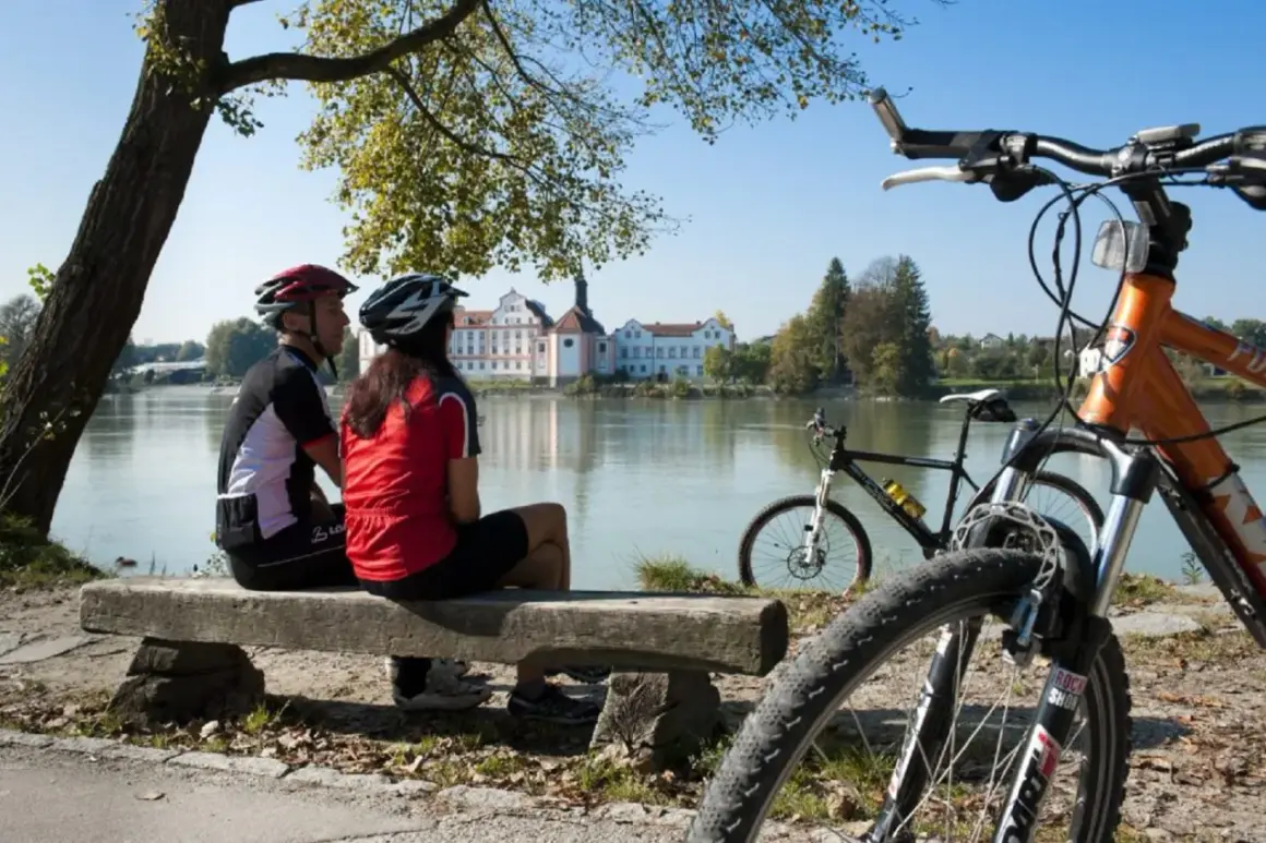 Para rowerzystów odpoczywa nad rzeką, podziwiając malownicze **trasy rowerowe Austrii** i pałac w oddali.