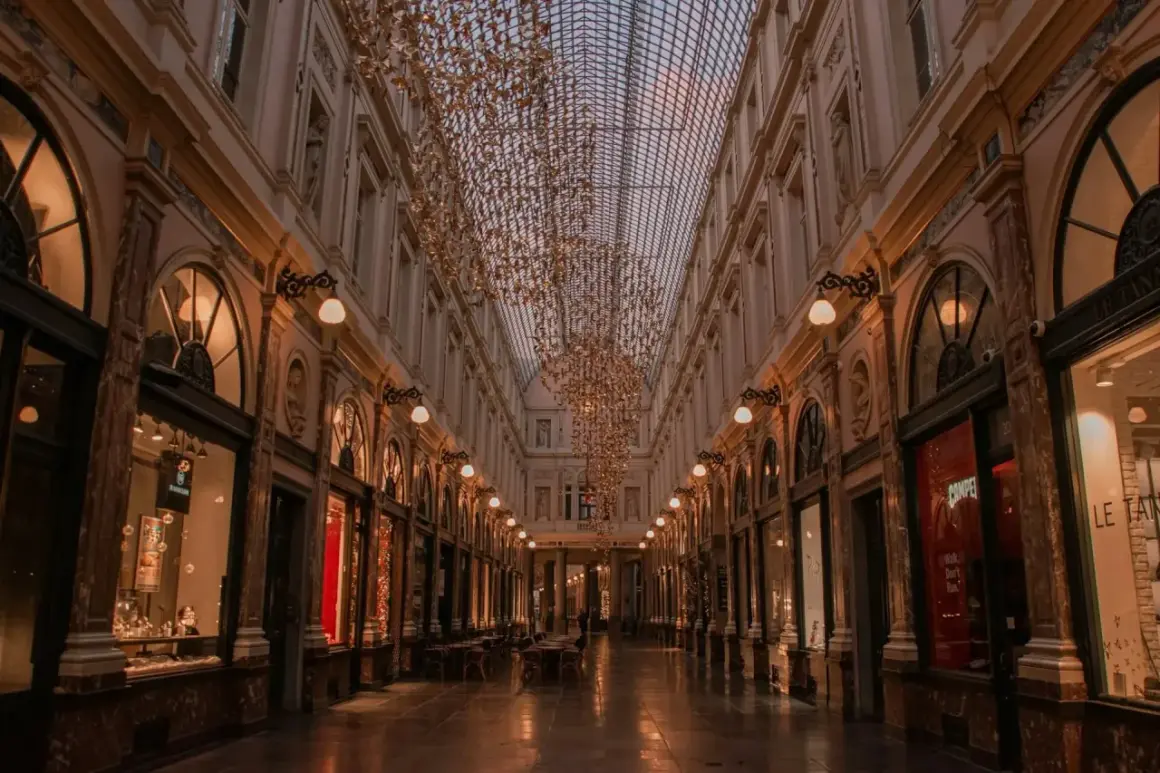 Elegancka galeria handlowa w Brukseli, z rzędem sklepów i kawiarni, ozdobiona złotymi, wiszącymi instalacjami. To urokliwe miejsce, które warto odwiedzić, zwiedzając brukselskie dzielnice.