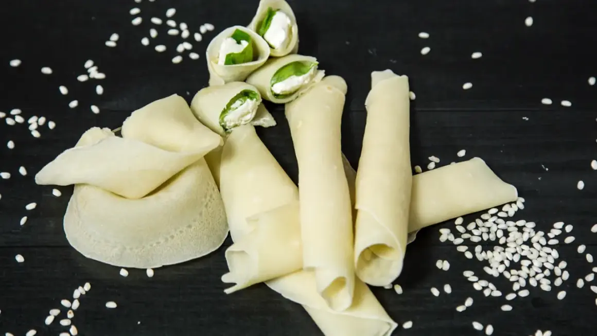 Rotolini di crepes con farina di riso, ripieni di formaggio fresco e basilico, sparsi su un tavolo scuro con semi di sesamo.
