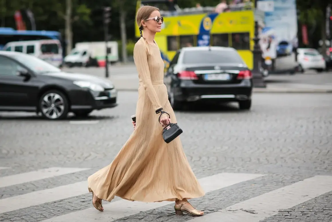 Elegancka, beżowa sukienka maxi, idealna na miejskie stylizacje. Kobieta w okularach przeciwsłonecznych i małej czarnej torebce przechodzi przez pasy.
