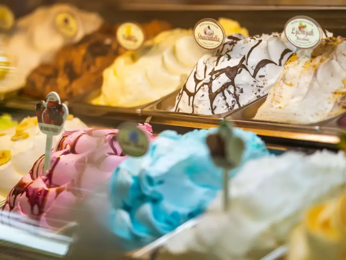 Verschiedene Eissorten in einer Auslage, darunter ein cremiges gelbes Eis, Stracciatella und ein fruchtiges Eis.