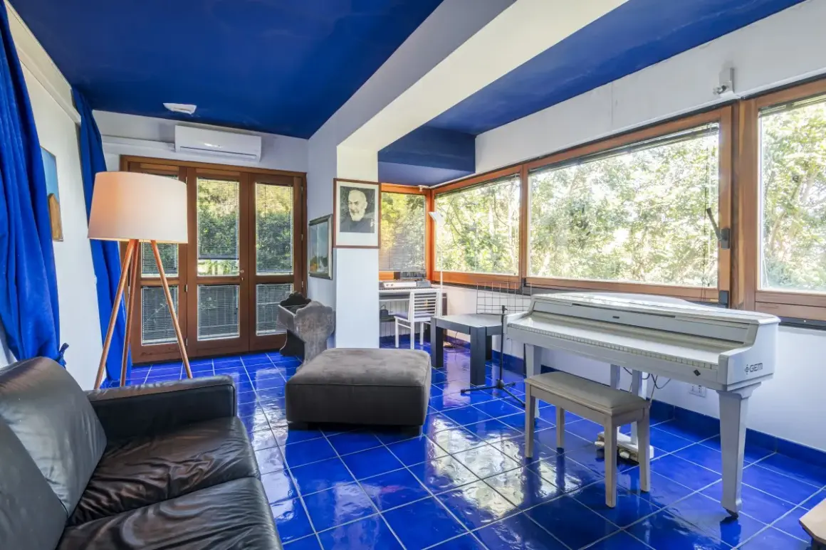 Interno con pavimento blu brillante, pianoforte bianco, divano nero e un ritratto di Padre Pio, come in testi lucio dalla la casa in riva al mare.