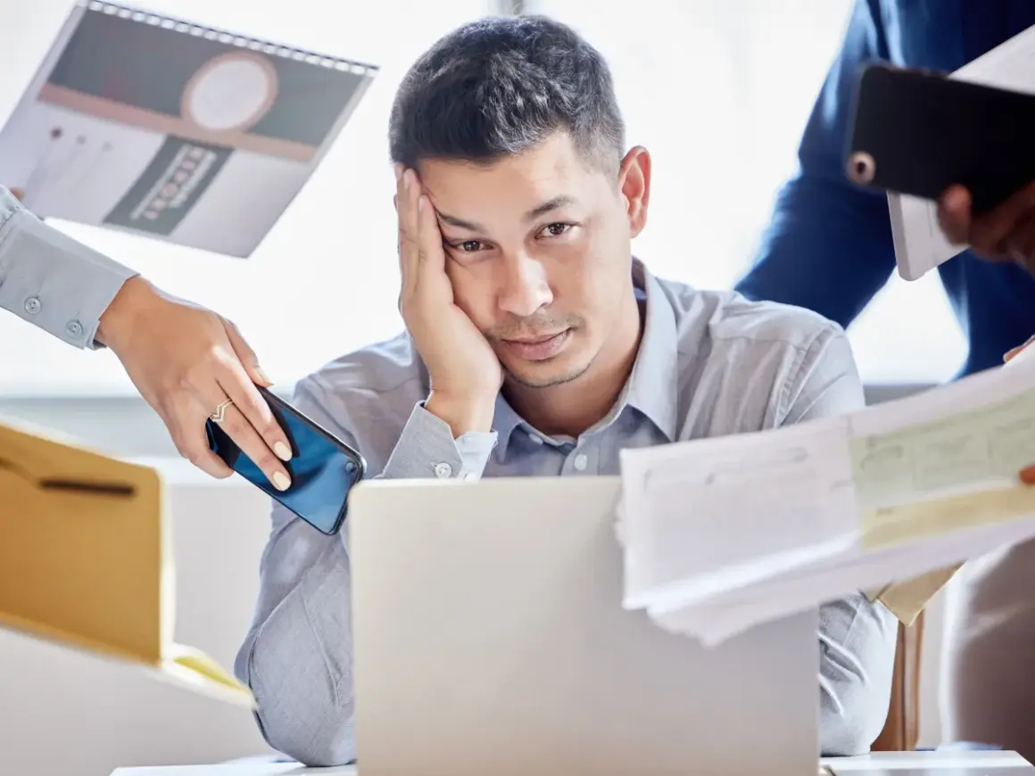 Zmęczony mężczyzna przy laptopie, otoczony dokumentami i telefonami. Czy to właśnie multitasking co to znaczy?