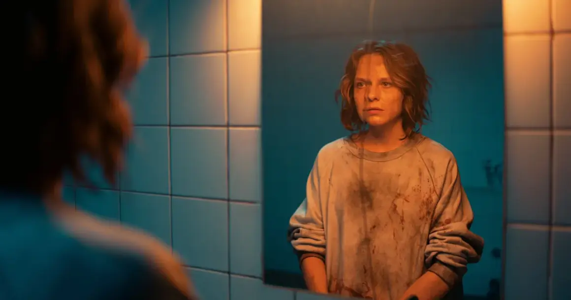 Mroczna scena z filmu "Morderczynie" - młoda kobieta w poplamionej bluzie patrzy w lustro, jej twarz wyraża niepokój.