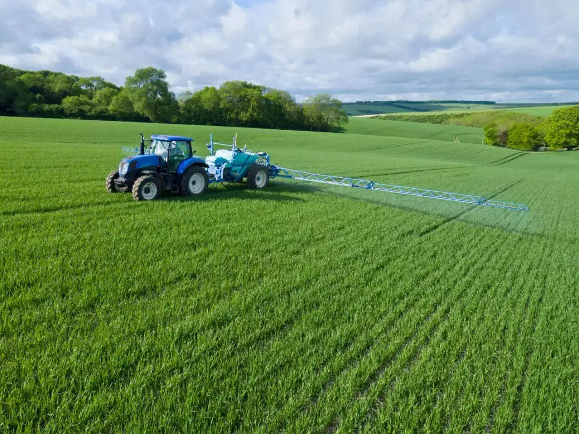 Niebieski traktor z opryskiwaczem na zielonym polu.