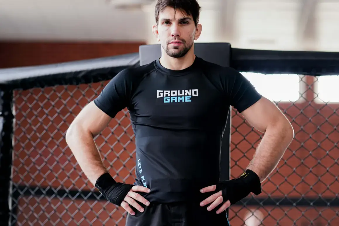 Mężczyzna w czarnym rashguardzie z napisem "GROUND GAME" stoi w klatce. Czy to jest to, co nazywamy rashguard co to?