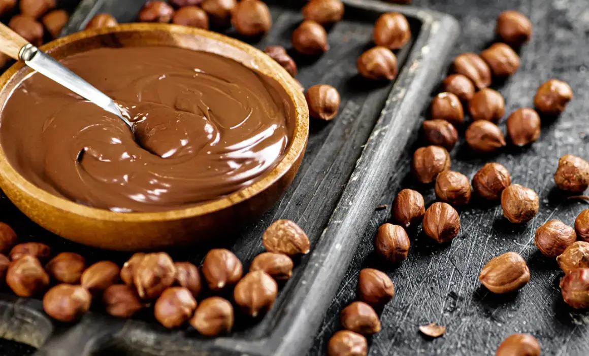 Schokocreme selber machen: Eine Schüssel voller cremiger Schokocreme neben gerösteten Haselnüssen auf einem Holzbrett.