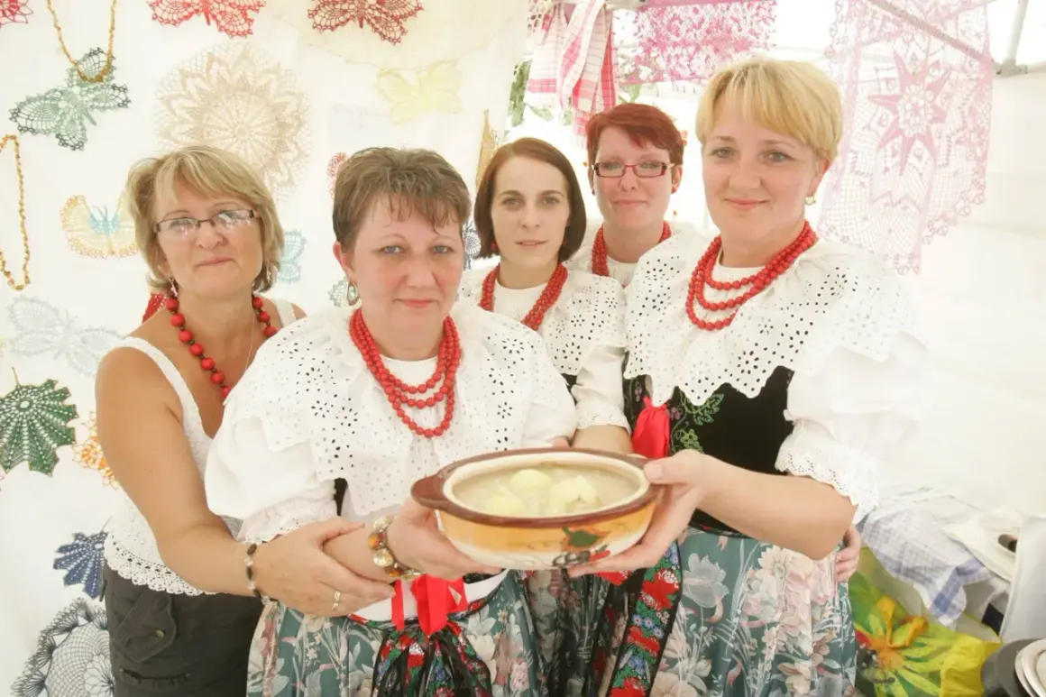 Pięć kobiet w tradycyjnych strojach ludowych prezentuje miskę z zupą. Jedna z nich trzyma ją w dłoniach. W tle widać ozdobne koronki, niczym z kukiełkowego teatru.