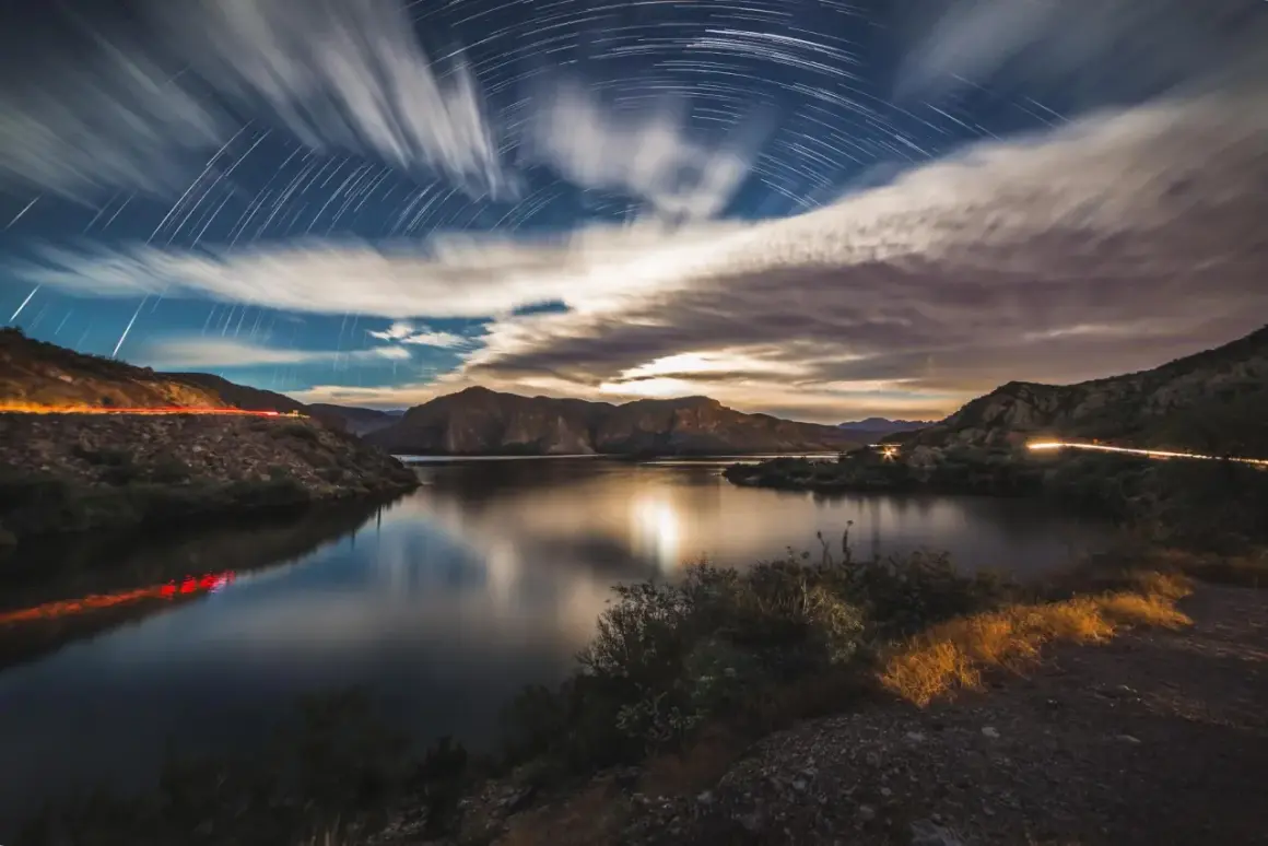 A surreal moon landscape with star trails arching over a calm lake and rugged mountains. Light trails from cars streak across the scene.