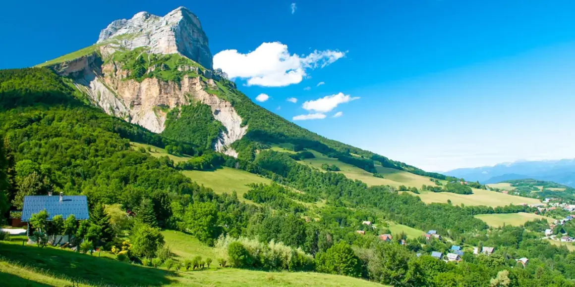 Vue panoramique d'une montagne imposante, le GR 965, dominant une vallée verdoyante parsemée de maisons. Le ciel bleu est parsemé de quelques nuages blancs.