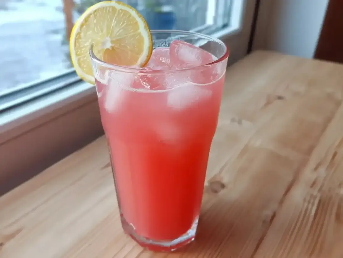 Ett glas rosa rabarberlemonad med is och en citronskiva, redo att avnjutas vid fönstret.