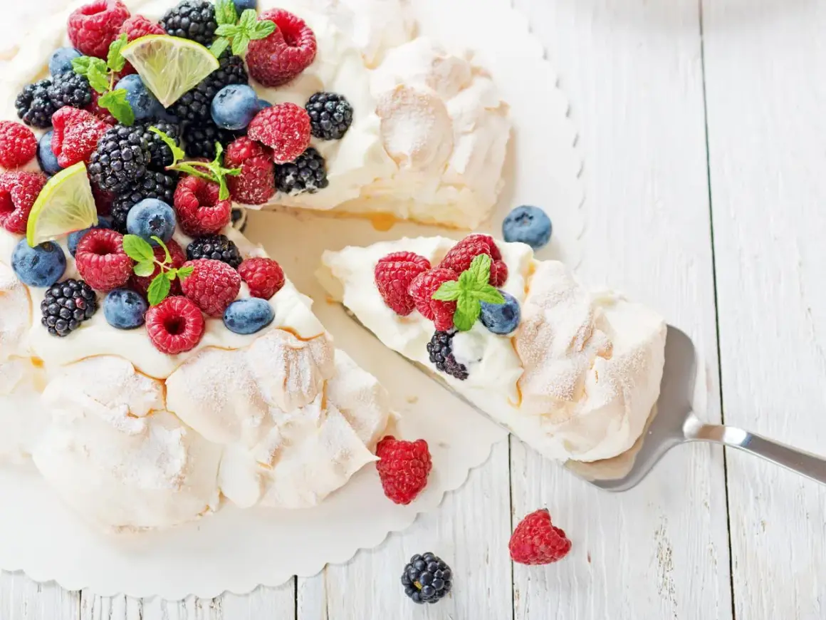 Beza Pavlova która zawsze wychodzi, udekorowana świeżymi malinami, borówkami, jeżynami i listkami mięty. Kawałek ciasta jest już nakładany.