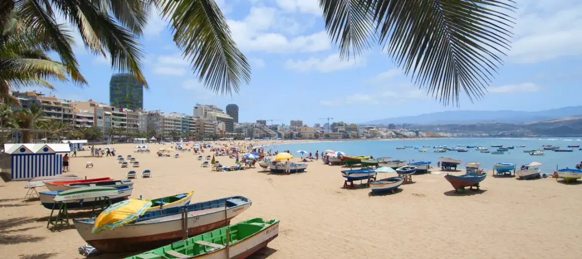 Solig strand i Las Palmas med palmer, båtar på sanden och en livlig stad i bakgrunden.