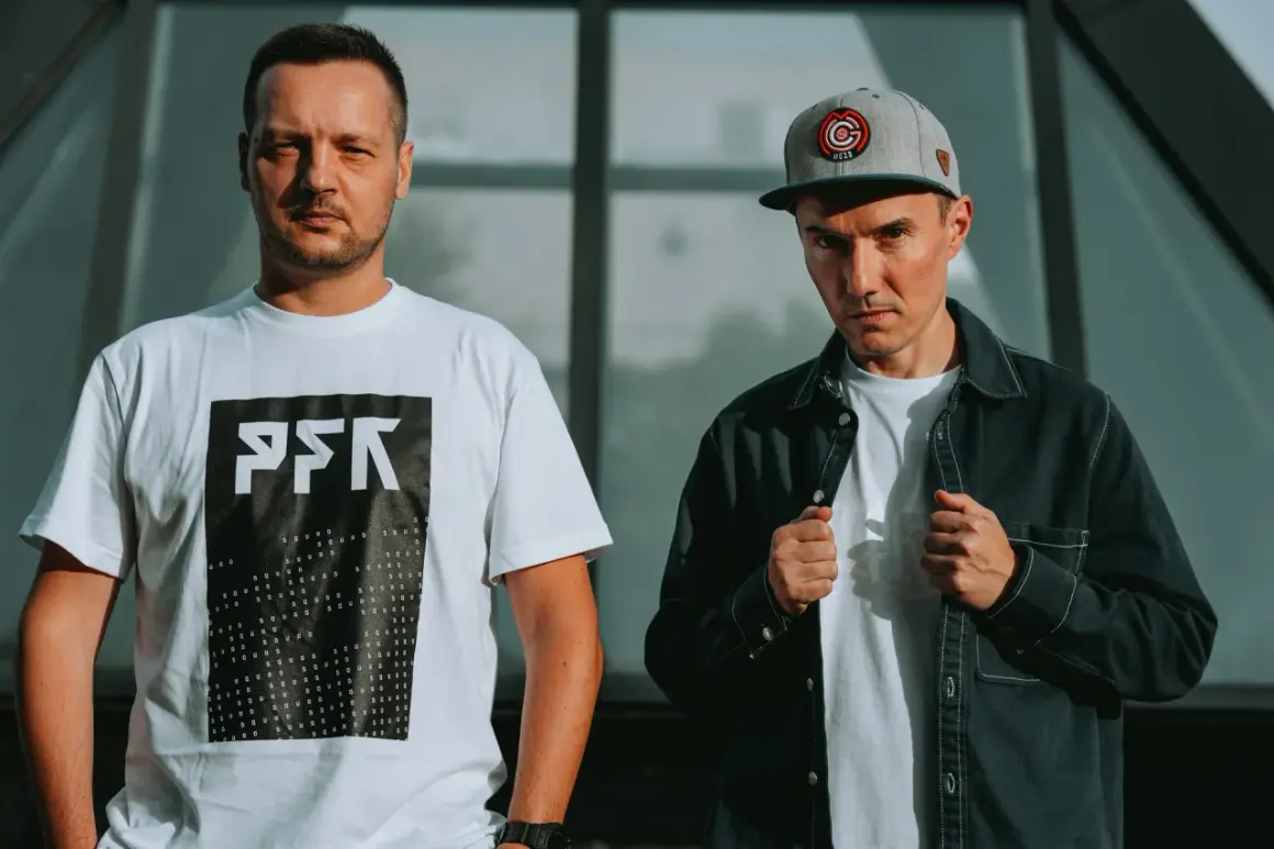 Dwóch członków Paktofoniki, jeden w białej koszulce z logo, drugi w czarnej kurtce i czapce z daszkiem.